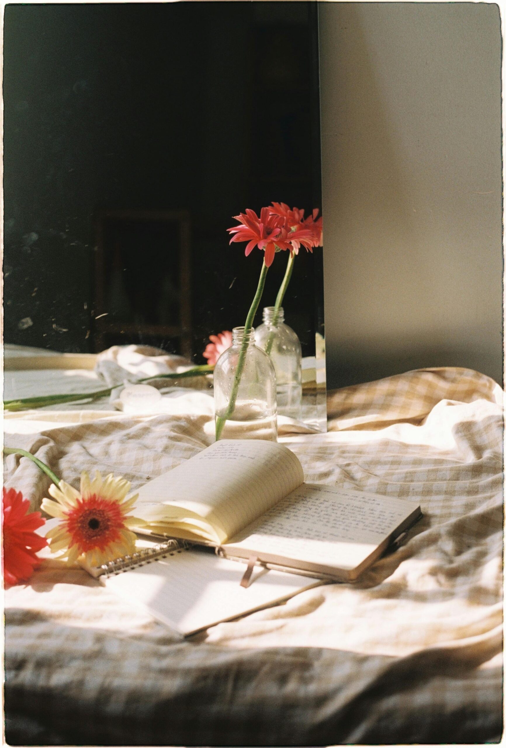 A cozy bedroom scene with gerbera flowers, a notebook, and sunlight streaming in.